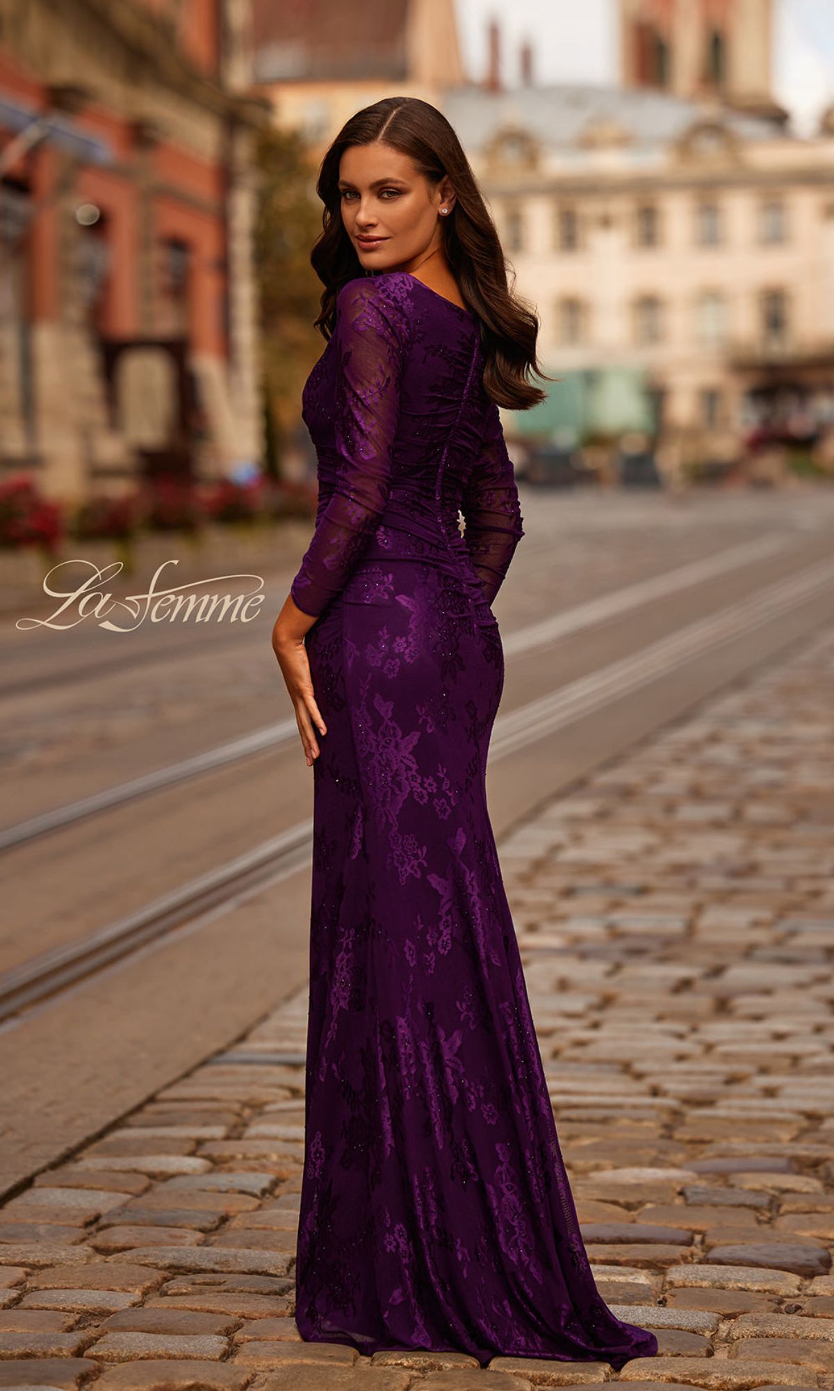 A woman in the Sheer-Sleeved Lace Formal Dress: La Femme 32491, a long-sleeved, floor-length purple lace gown with sheer sleeves, stands on a cobblestone street and looks over her shoulder.